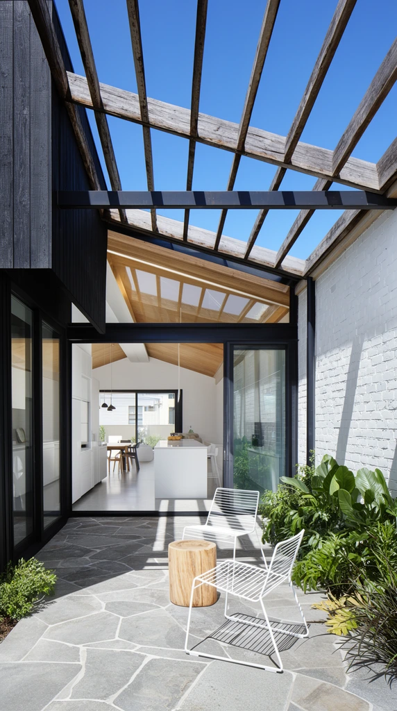 A modern courtyard transitions seamlessly into a bright open-plan interior beneath a clear blue sky. Two white wireframe chairs and a wooden stump table rest on irregular grey stone paving beside lush greenery. An overhead pergola of weathered wooden beams and dark metal frames casts strong diagonal shadows across the patio. The striking architecture contrasts black vertical timber cladding on the left with white painted brick on the right. Wide-open glass doors frame a sleek white kitchen island, dining space, and pitched timber ceiling. Captured in crisp architectural photography with high-contrast daylight.