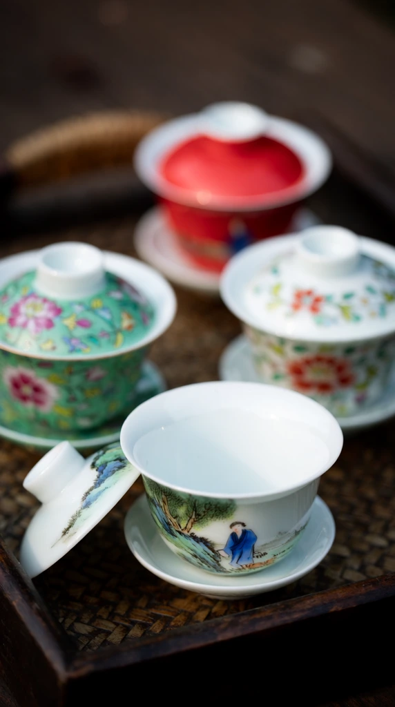 five traditional porcelain teacups on a dark woven rattan tray, open white teacup in sharp foreground focus featuring a painted landscape with green trees and a blue-robed figure, matching lid resting nearby, three closed middle-ground teacups decorated with vibrant floral patterns in green pink and red, blurred bright red teacup in the background, extremely shallow depth of field, smooth bokeh, soft natural daylight highlighting the glossy porcelain, macro still life photography, dark wooden setting