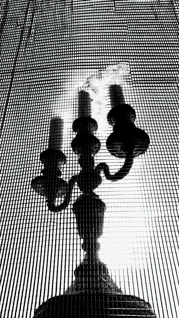 low-angle worm's-eye view of a classic metal candelabra with lit candles and wisps of smoke seen through a dense industrial metal grate, high-contrast black and white, stark binary aesthetic, heavy vertical and horizontal line screen, gritty halftone texture, silhouette-focused composition, dark atmospheric mood, minimalist graphic design style, sharp grid overlay, monochromatic, experimental film photography, brutalist influence, centered subject, high-impact visual.
