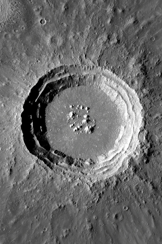 A massive circular impact crater dominates a barren, grayscale planetary surface from a top-down orbital perspective. Steep, terraced inner walls step down to a flat basin containing a cluster of jagged central peaks. Harsh low-angle lighting casts deep black shadows across the left inner rim, sharply defining the intricate topographical relief. The surrounding desolate landscape is heavily textured, scarred with numerous smaller secondary craters, radiating ejecta ridges, and coarse rocky debris. Stark high-contrast shading highlights the rugged geology with high-resolution photographic clarity.