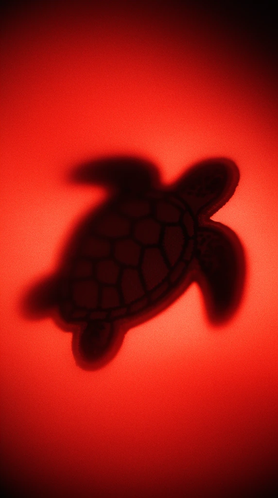 A dark, heavily blurred silhouette of a stitched sea turtle patch angled toward the upper right, suspended within a luminous, highly saturated crimson red environment. The form of the sea turtle patch is obscured by extreme soft focus, appearing as a shadowy, indistinct mass with smooth, bleeding edges that swallow all fine thread details. The intense red background features a heavy, hazy atmospheric diffusion, glowing brightest in the center directly behind the subject. A pronounced deep vignette frames the composition, heavily darkening the outer corners and concentrating the backlit illumination inward. The entire composition is enveloped in a strong, coarse grain and thick optical noise, creating a stark, high-contrast separation between the deep black shadow of the angled turtle and the glowing, monochromatic red void.