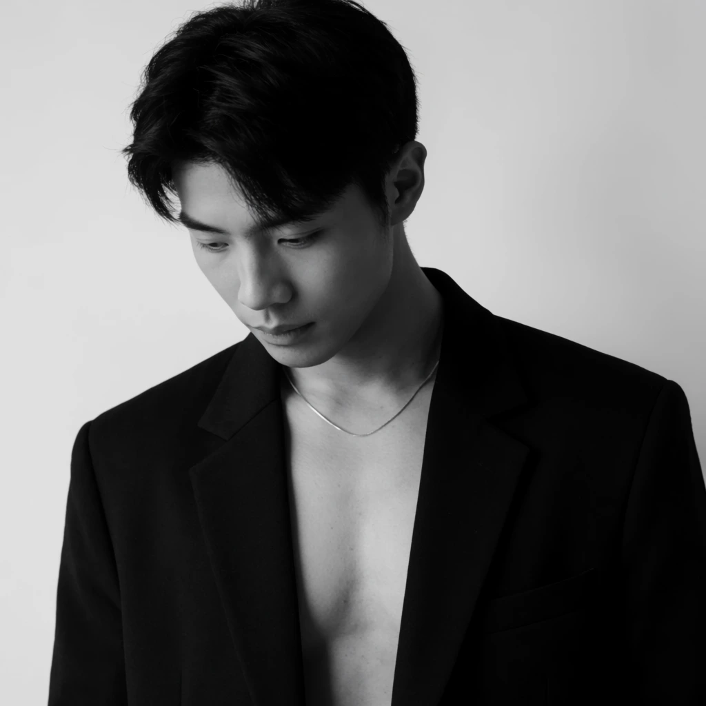 black and white studio portrait, young Asian man looking down, dark hair, open black blazer over bare chest, thin silver chain, soft directional lighting, plain background, minimalist fashion photography