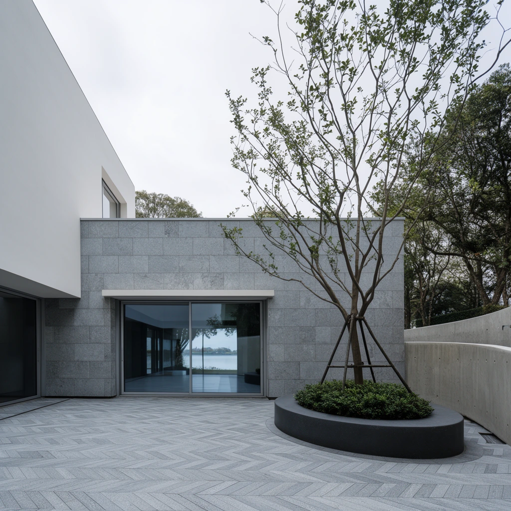 modern minimalist courtyard, chevron-patterned grey pavement, building facade clad in large grey stone tiles, sliding glass doors reflecting distant water, adjacent smooth white architectural volume, circular dark grey planter with a slender guy-wired tree and low green shrubs, curved concrete retaining wall on the right, mature background trees, overcast sky, architectural photography, wide-angle perspective, soft diffused daylight, clean geometric composition