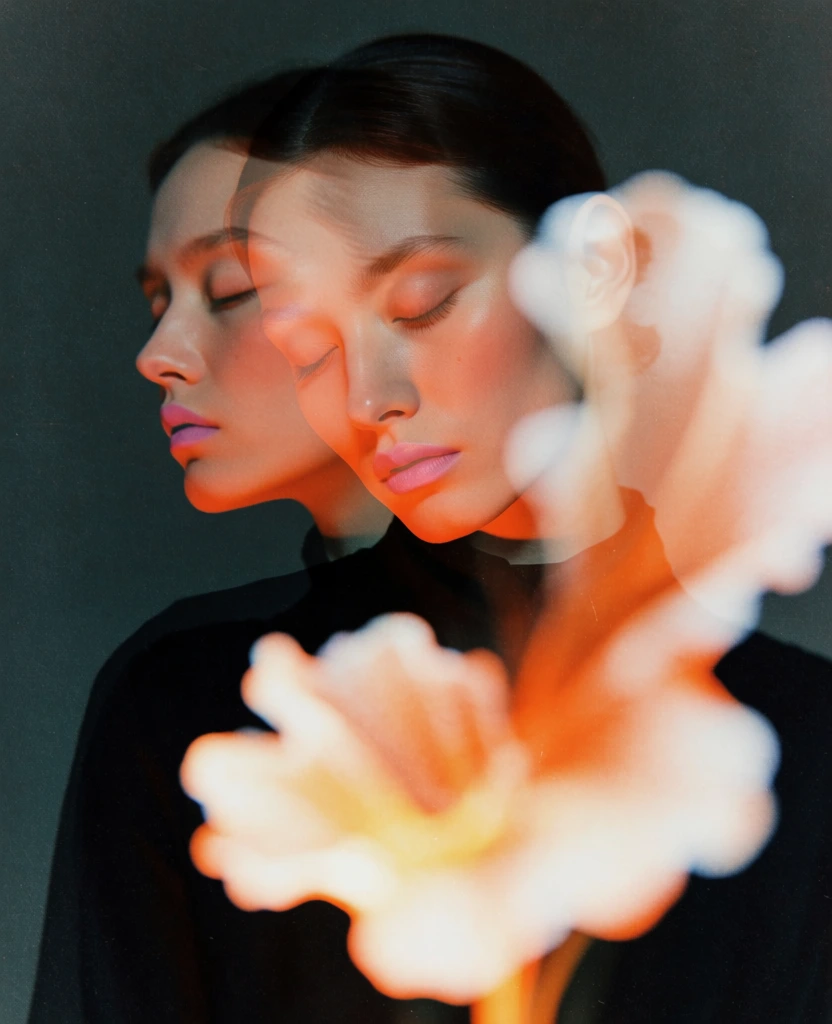 A double-exposure-style portrait features two overlapping, translucent images of a person's face against a dark, grainy background, accompanied by a glowing, out-of-focus flower in the foreground. The central subject is a person with pale skin and dark hair pulled back, appearing in two distinct poses; one face is seen in a crisp profile looking toward the left, while the other is tilted forward and turned slightly to the right, overlapping the first. Both faces are adorned with warm peach-toned eyeshadow and a matching soft lip color, with light catching the high points of the cheekbones and forehead. The person wears a dark, black garment that merges with the shadows of the lower frame. In the immediate foreground, a large, blurry, and brightly illuminated flower-like shape in shades of white and orange glows intensely, partially obscuring the lower half of the subject. The background is a uniform, dark teal-grey with a heavy, tactile grain throughout the entire composition. The artistic style utilizes a soft-focus technique and low-light conditions, creating a dreamy, ethereal atmosphere with a color palette centered on deep cool tones contrasted by warm, luminous highlights.
