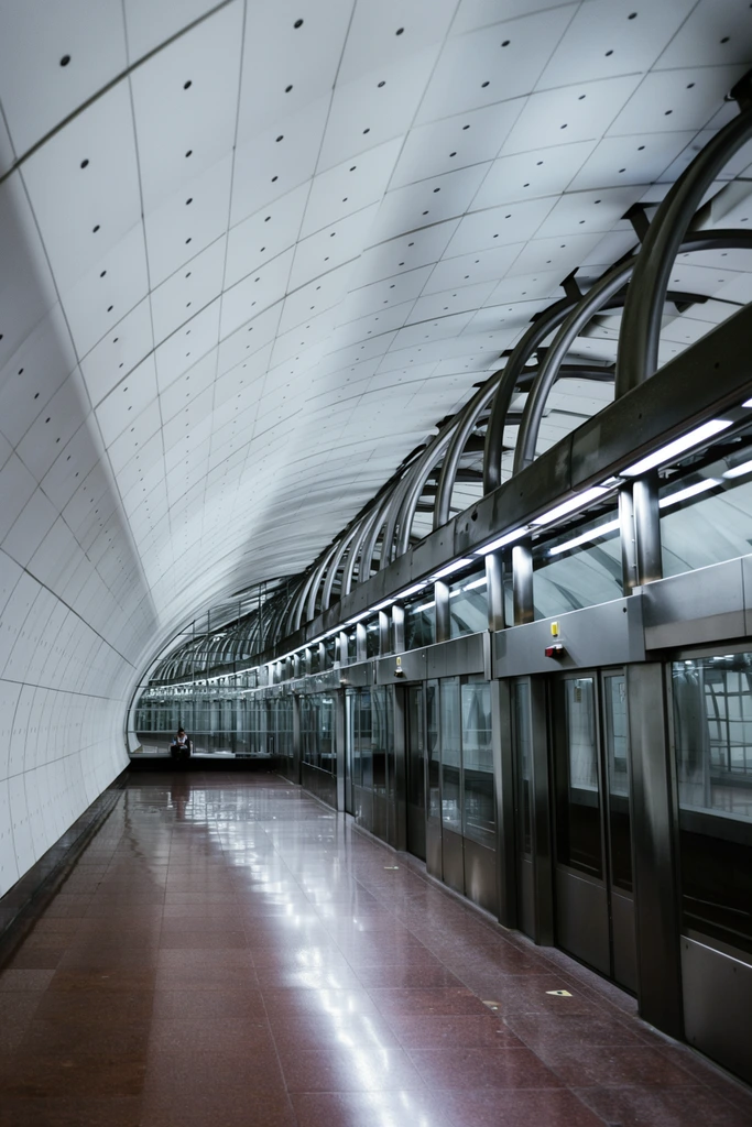 A subterranean transit platform curves deeply into the background beneath a vaulted, white paneled ceiling punctuated by small circular details. Along the right, a continuous barrier of glass and brushed steel automated doors is framed by heavy curved metal beams and bright linear lights. The polished, reddish-brown tiled floor sharply reflects the cool, sterile illumination, enhancing the sweeping architectural perspective. A solitary figure sits faintly visible behind the distant glass within the sleek, geometric, wide-angle interior.