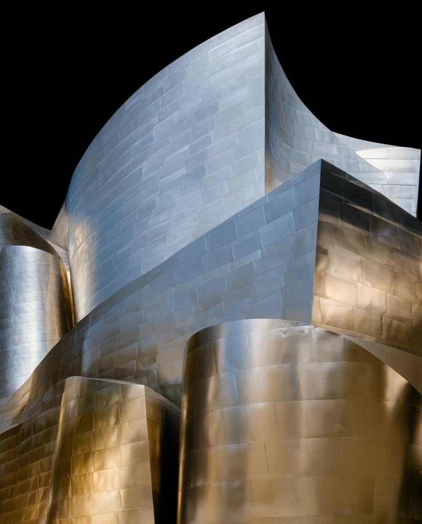 Sweeping deconstructivist architectural forms clad in rectangular brushed metal panels surge upward against a stark, pitch-black background. The massive structure features overlapping, wave-like metallic facades and smooth cylindrical volumes that contrast with sharp, angular protrusions. Precise grid lines of the cladding seams map the undulating exterior. Dramatic lighting creates striking reflections across the surface, casting warm golden-bronze highlights on the lower and rightward curves that transition into cool, silvery-blue tones on the towering upper sections. Shot from a low angle, the composition emphasizes the fluid, organic geometry and monumental scale of the reflective metallic architecture isolated within a dark void.
