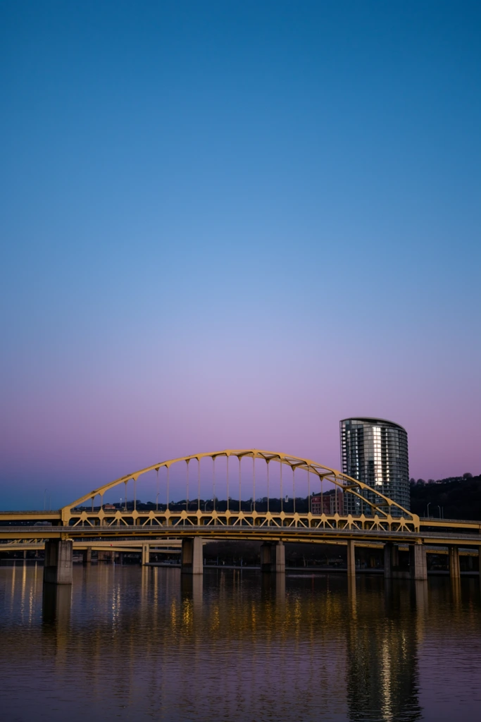 yellow arch bridge spanning a calm river at dusk, vast clear sky with a smooth color gradient from deep blue to soft magenta at the horizon, vertical suspension cables, concrete bridge piers, golden light reflections shimmering on the dark water, modern curved high-rise building with illuminated windows in the background, dark silhouette of distant rolling hills, expansive landscape photography, serene atmosphere, sky dominating the upper composition