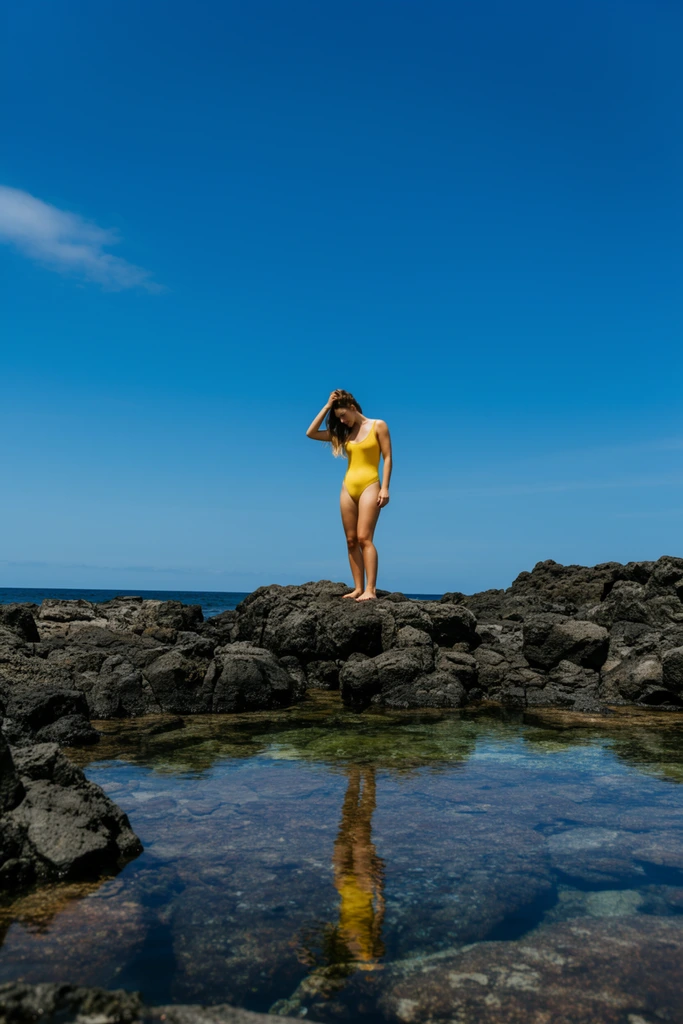 Woman in a bright yellow one-piece swimsuit standing atop jagged dark volcanic rocks, touching her hair while looking down, calm clear tide pool in the foreground capturing her reflection, deep blue ocean horizon, expansive vivid blue sky with a faint wispy cloud, bright midday sunlight, stark shadows on textured stone, wide landscape shot, low angle