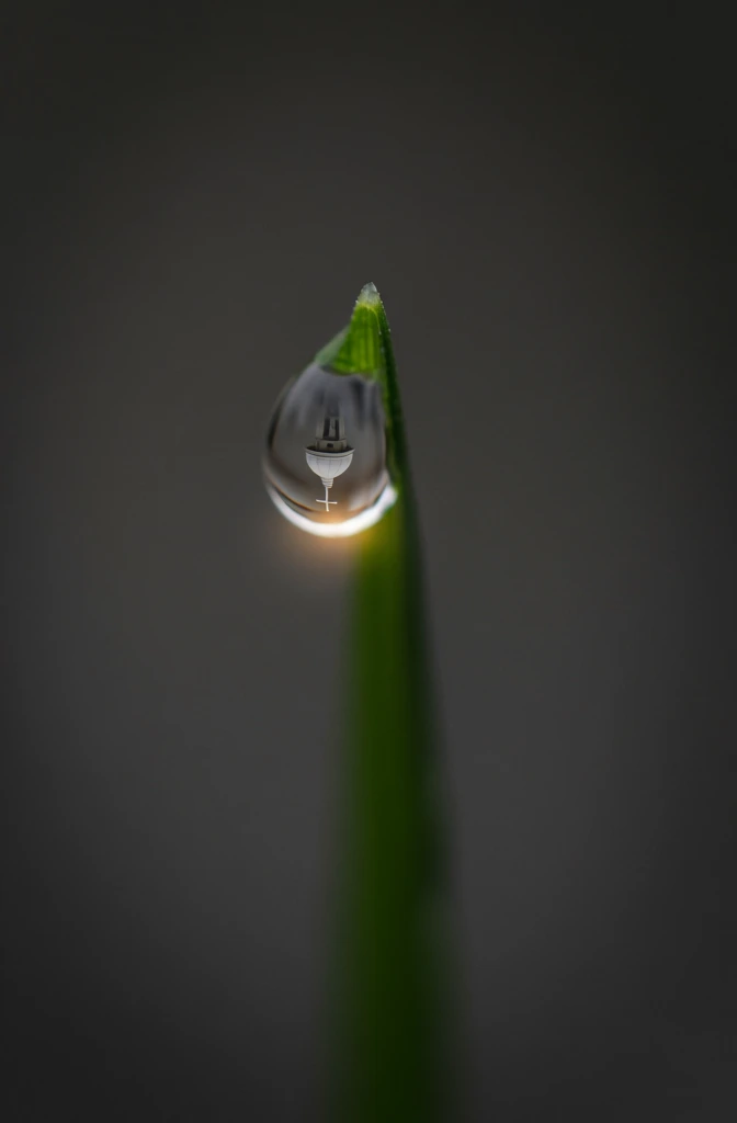 macro photography of a single crystalline water drop hanging from a sharp green grass blade, minimalist dark grey background, shallow depth of field, spherical refraction inside the droplet showing an inverted view of one dome with a grid pattern topped by one cross, warm golden hour light catching the bottom edge of the drop with a subtle sun flare, high contrast, clean vertical composition, crisp focus on the droplet, elegant architectural macro aesthetic