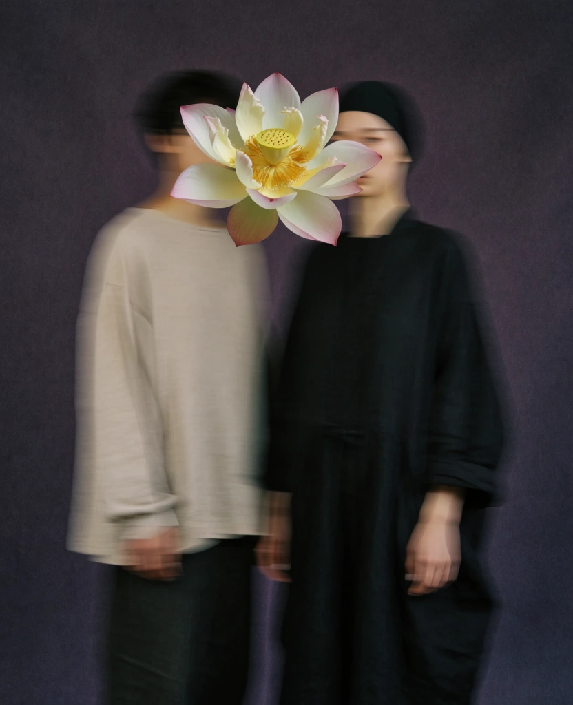 An artistic editorial advertisement features two blurred figures standing side-by-side with a large lotus flower obscuring their faces against a grainy, desaturated background. Two people stand centrally; the figure on the left wears an off-white, voluminous long-sleeve garment and dark trousers, while the figure on the right is dressed entirely in black, including a head covering that reveals only the eyes. A large, blooming pale-yellow lotus flower with a light pink center is positioned in the foreground, overlapping the head area of both figures. The humans are captured with a heavy motion blur effect, creating soft, streaked outlines that suggest movement. The backdrop is a solid, textured, dark purple-gray surface. At the top center, "THE NORTH FACE" logo in white is placed next to the "Spiber" logo, which consists of a diamond-like icon and the brand name. Across the bottom third, the phrase "Utopian Uniform" is written in a large, bold, white, rounded font. Small green katakana characters are positioned directly above several letters of the main title: "ユ" above the "U", "ト" above the "t", "ピ" above the "o", "ア" above the "p", "ン" above the "n", "ユ" above the "U", "ニ" above the "n", "ホ" above the "i", "ー" above the "r", and "ム" above the "m". At the very bottom, a row of brand names is listed in small, white, sans-serif capital letters: "Goldwin", "THE NORTH FACE", "nanamica", "NEUTRALWORKS.", and "THE NORTH FACE Purple Label". The composition is symmetrical and centered, using a low-key lighting setup that creates deep shadows and a moody atmosphere. The overall aesthetic is characterized by a heavy film grain and a desaturated color palette of muted purples, grays, and creams, utilizing a straight-on medium shot perspective.