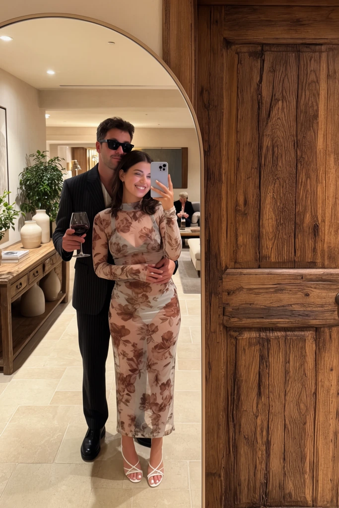 A couple takes a mirror selfie in an arched floor mirror positioned next to a heavy rustic wooden door. A smiling woman in a form-fitting, sheer, long-sleeved dress with a brown floral print holds a smartphone, wearing white strappy heels. A man in a dark pinstripe suit and black sunglasses stands behind her, embracing her waist with one arm while holding a glass of red wine in his other hand. The mirror reflection reveals a warm indoor lounge with a tiled floor, a wooden console table displaying vases and a potted plant, and a distant figure seated on a sofa. Soft, warm indoor lighting illuminates the couple, highlighting the thick wood grain of the adjacent door and the semi-transparent texture of the floral dress.