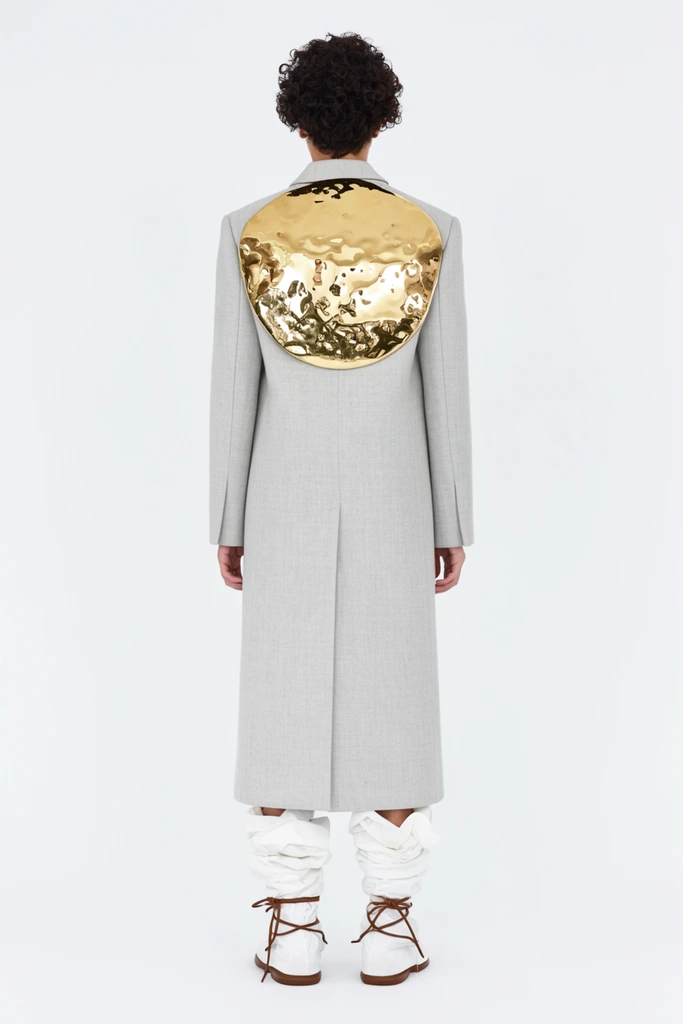 rear view of a person in a long light grey tailored coat, large reflective hammered gold plate attached to the upper back, short dark curly hair, wrapped white fabric boots tied with brown laces over brown shoes, minimalist stark white background, avant-garde fashion photography, bright even studio lighting