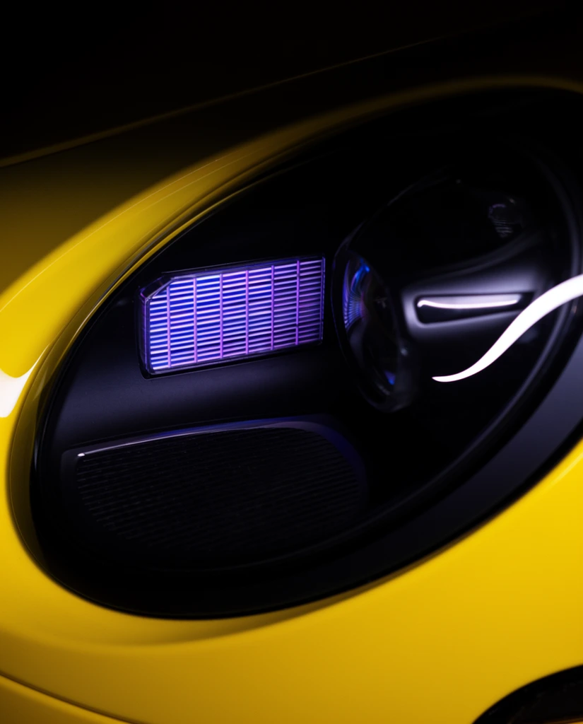 A close-up macro photograph of a vivid yellow sports car headlight assembly emerging from deep black shadows. The glossy, curved yellow body panel encases a large, dark circular headlamp. Inside the glass housing, an advanced lighting system displays a glowing purple-blue rectangular grid of thin horizontal LED bars. Beside this grid rests a dark cylindrical projector lens highlighted by a sharp, curved white glowing accent strip. Tightly cropped and highly contrasting, the composition emphasizes sleek automotive design, blending smooth painted exterior curves with complex internal optical technology.