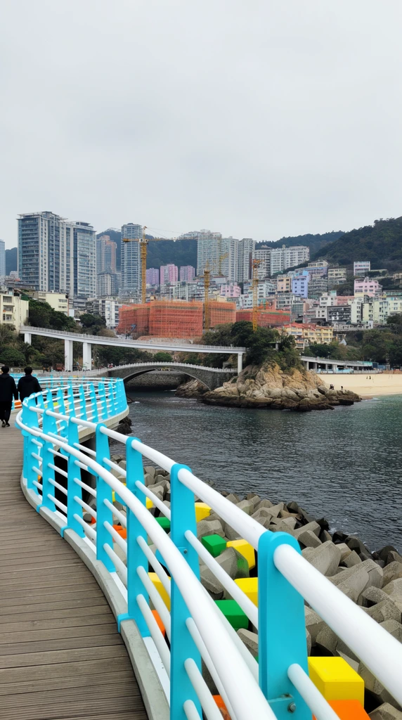 A curved wooden pedestrian boardwalk with bright cyan and white metal railings sweeps over dark coastal waters toward a densely built, mountainous city. Two figures in dark jackets walk away along the left foreground. Below, grey concrete tetrapods line the rocky shoreline, accented by solitary bright yellow, green, and orange blocks. Across the water, an arched stone bridge and a slender white overpass connect a rocky islet to a sandy crescent beach. The sprawling urban backdrop features modern high-rise towers, pastel buildings, and construction sites draped in orange mesh with tall yellow cranes. Lush forested hills rise immediately behind the dense architecture beneath a flat, overcast grey sky. The wide-angle perspective and diffused daylight emphasize the bright industrial colors against the muted coastal urban landscape.