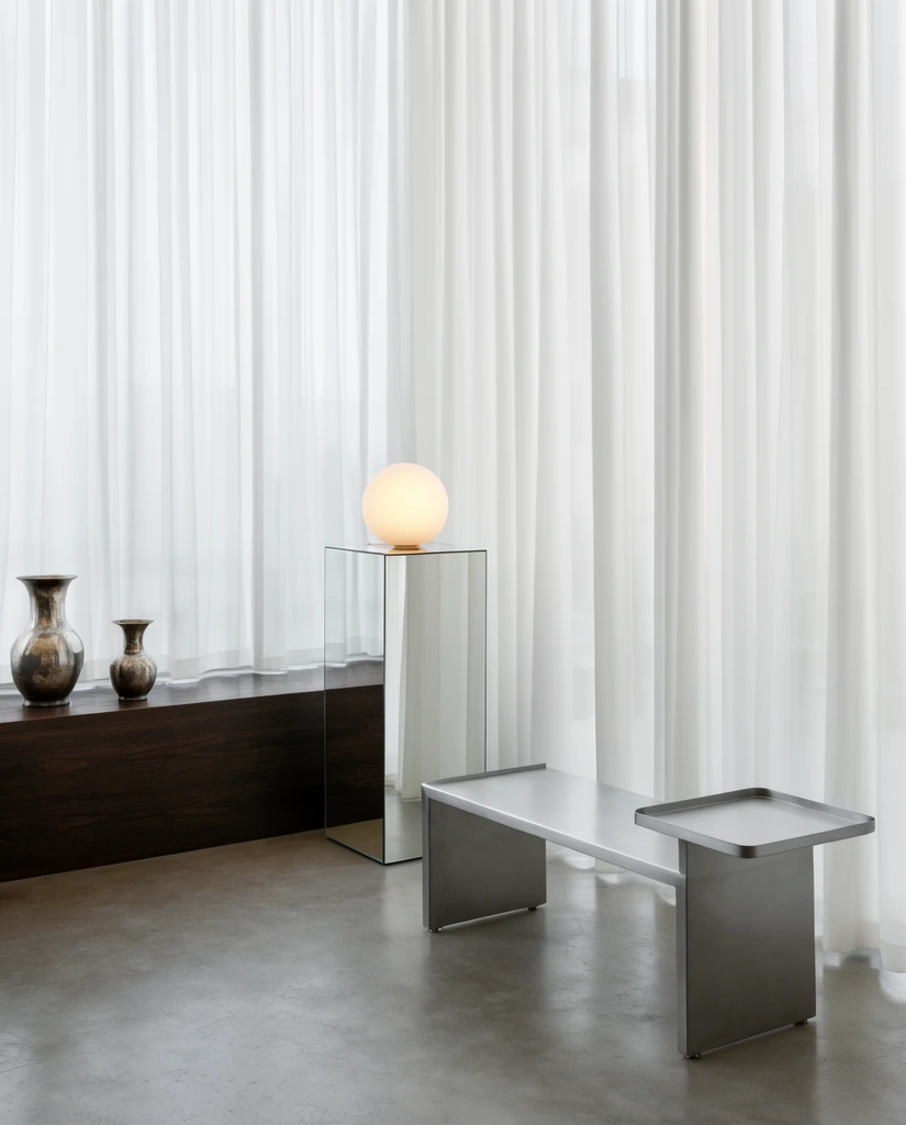 flowing sheer white curtains, minimalist interior, dark wood plinth with two metallic vases, mirrored pedestal with glowing spherical lamp, metal bench with attached side table, polished concrete floor, soft diffused daylight, architectural photography