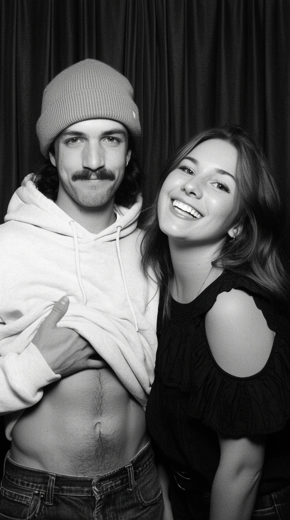 black and white photobooth portrait, playful man and woman against dark pleated curtains, man on left wearing light hoodie and beanie lifting shirt to expose bare stomach, sporting a mustache and subtle smirk, woman on right leaning in with bright joyful smile wearing dark ruffled cold-shoulder top, direct flash, grainy texture