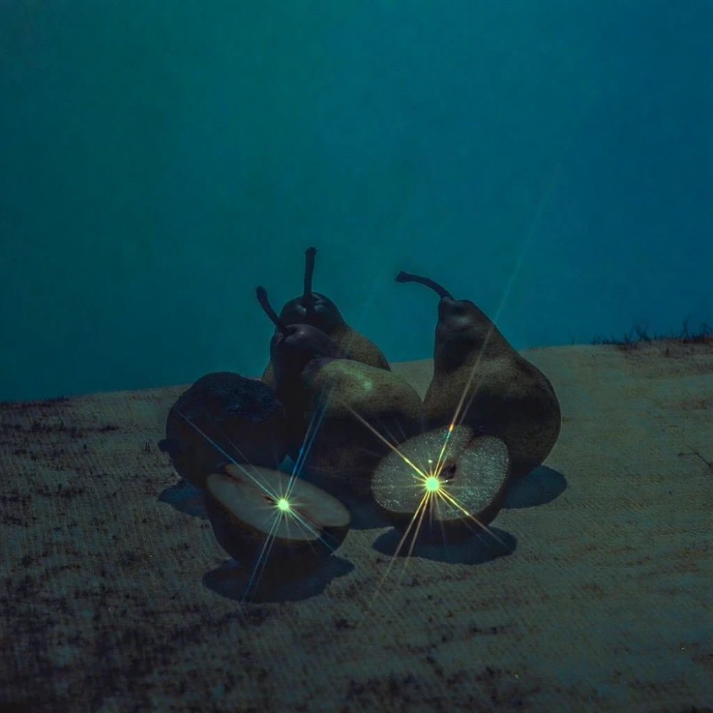 a cluster of whole pears and round fruits with two halved fruits in the foreground resting on a sharp diagonal stripe, monochromatic nautical teal and cyan color grade, underexposed low-key lighting, sharp 8-point starburst lens flares emerging from the fruit cores, heavy analog film grain, crushed blacks with deep blue-green midtones, moody cinematic still life, ethereal bioluminescent glow, lo-fi aesthetic, soft focus edges, high-contrast chiaroscuro, surreal liminal atmosphere.