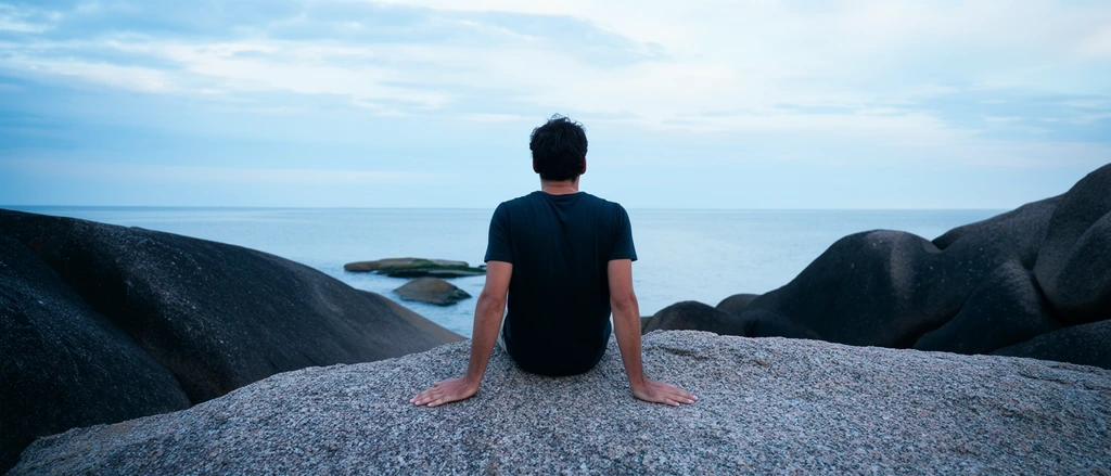 rear view of a dark-haired man sitting on a massive speckled grey boulder facing a calm ocean, wearing a dark short-sleeved t-shirt, hands resting wide on the rough rock surface, surrounded by smooth dark granite formations, tranquil sea extending to a flat horizon, distant partially submerged rocks, overcast sky with soft blue clouds, wide landscape photography, cool color palette, serene atmosphere, diffused natural daylight
