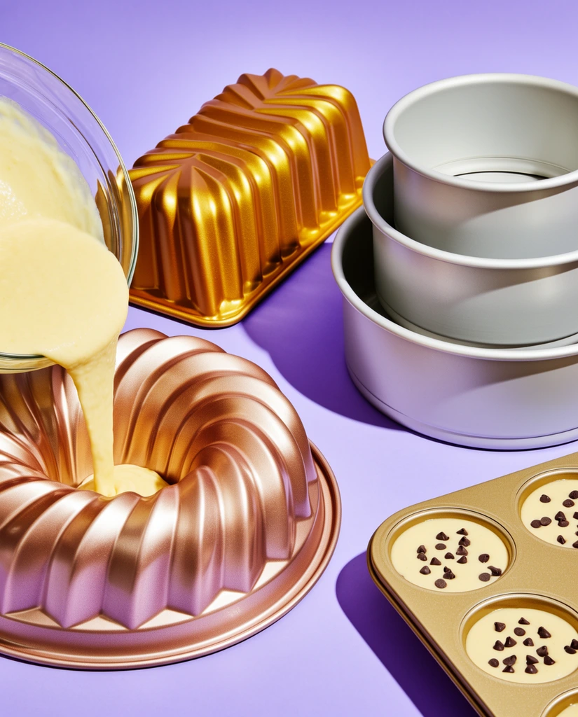 Thick, pale yellow batter pours from a clear glass bowl into a metallic rose-gold, fluted ring pan on the left. Beside it, an inverted, deeply ribbed rectangular loaf pan gleams with a bright gold finish. To the right, three smooth, matte light grey round cake pans are stacked in descending size. In the lower right corner, a gold-toned muffin tin contains pale batter sprinkled with dark chocolate chips. The meticulously arranged baking tools rest on a seamless, solid lavender surface. Hard, directional studio lighting casts sharp, deep purple drop shadows to the left of each object, highlighting the contrast between the highly reflective metallic ridges and the smooth matte surfaces. The still-life composition features a high-angle perspective characteristic of bold, contemporary commercial product photography.