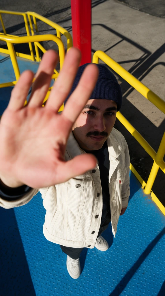 A high-angle, wide-lens photograph looking down at a young man with a mustache, wearing a navy beanie and cream corduroy jacket. He reaches an out-of-focus, open hand directly toward the lens, dominating the foreground. Intense sunlight casts a sharp shadow of his hand across his face. He stands on a vibrant blue diamond-plate platform bordered by bold yellow tubular railings and a red post. Dark, crisp shadows stretch across the surrounding concrete, accentuating the high-contrast lighting and striking primary color palette of the industrial environment.