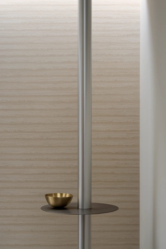vertical brushed silver pole against a beige wall with horizontal textured striations, small polished brass bowl resting on a thin circular dark metal disc attached near the base, smooth white wall edge framing the right, soft top-down lighting, minimalist contemporary interior design, serene aesthetic, photorealistic