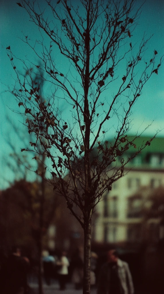 slender bare tree with sparse dry leaves and intricate dark branches in sharp foreground focus, vivid teal sky backdrop, heavily blurred urban background, out-of-focus distant building with a green roof, indistinct blurred walking figures at the base, vintage film photography, heavy grain, cross-processed cyan and deep brown color palette, extreme shallow depth of field, moody atmospheric lighting, high contrast silhouette effect