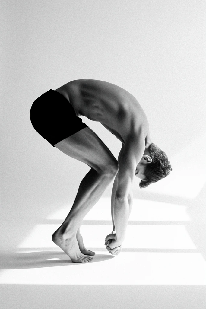 A stark black and white studio photograph of a shirtless man bending deeply forward at the waist in profile view. Wearing dark fitted shorts, he balances barefoot on the balls of his feet with arms extended straight down, hands tightly clasped near the floor. His head is tucked inward against a seamless white backdrop with prominent film grain. Intense directional light from the right casts sharp geometric shadows across the pristine floor, creating dramatic high-contrast lighting that sculpts the taut musculature of his arched back and legs.