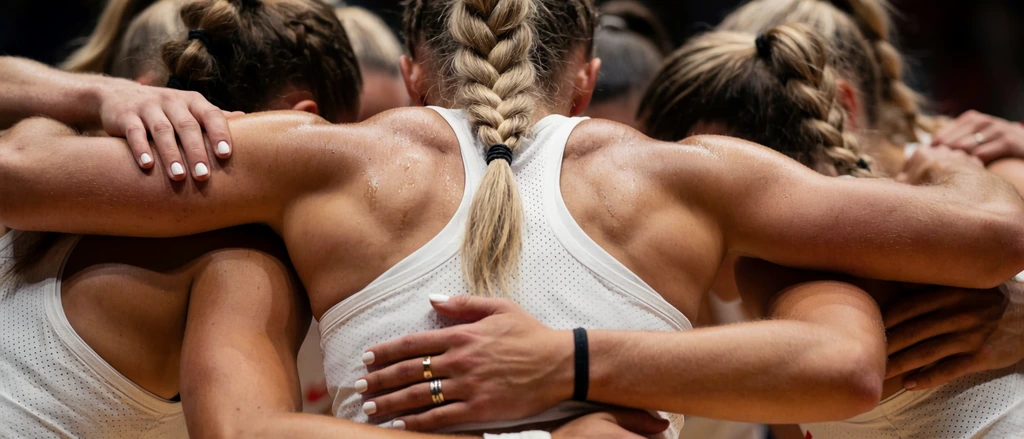 A tight close-up of female athletes locked in an intense team huddle. The central figure, viewed from behind, features muscular, sweat-glistening shoulders and a long blonde braided ponytail secured with black hair ties. She wears a white perforated athletic tank top. Teammates surround her in a dense embrace, their tanned arms interlocking across shoulders and waists. One visible hand resting on a waist displays white nail polish, a gold band, a silver ring, and a black wristband. Dramatic overhead lighting emphasizes the damp skin, the intricate texture of the hair, and the physical exertion of the group. The background is heavily blurred in dark, warm tones with a shallow depth of field. This sports photography composition highlights the emotional weight and interlocking geometry of the unified team.