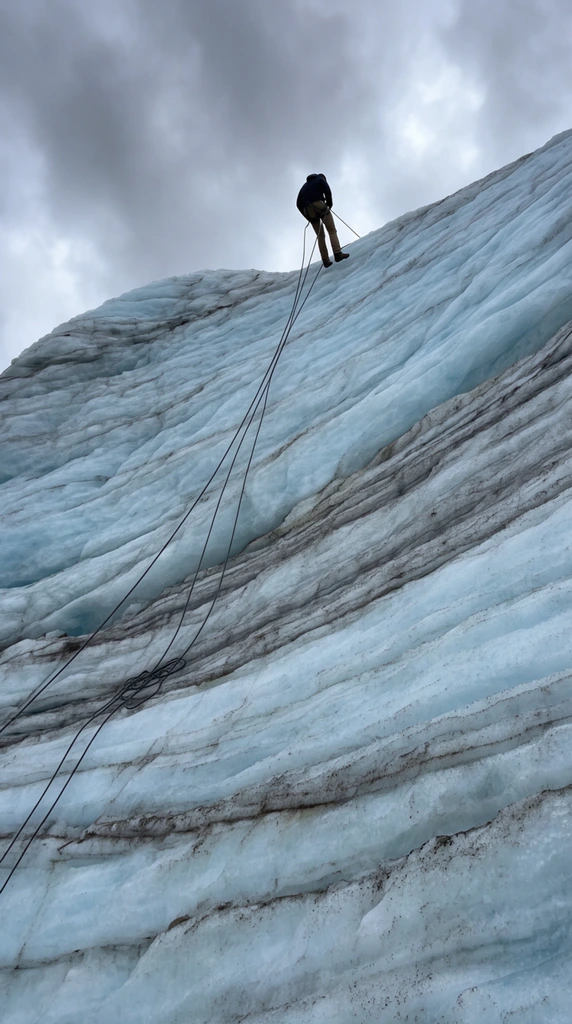 A solitary climber rappels down a massive, striated glacier under an overcast sky. Photographed from a dramatic low angle, the small silhouetted figure in a dark jacket and tan pants descends a steep, pale blue ice wall. Dark climbing ropes trail down the rugged face, intersecting prominent horizontal bands of grey dirt and deep textural ridges. The sweeping scale of the layered ice dominates the composition, emphasizing the climber perched near the top crest against dense grey clouds.