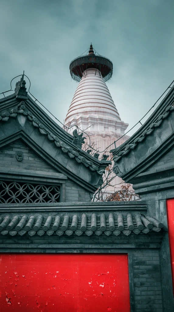 Traditional dark gray tiled roofs and vibrant red walls frame a towering white stupa from a low-angle perspective. The foreground showcases layered cylindrical roof tiles above a gray brick wall featuring geometric circular latticework. Below this, a solid panel of distressed red paint with slight peeling offers a stark color contrast. Thin dark wires and sparse bare branches intersect the roofline. In the background, the massive white conical structure displays horizontal ridged banding and an ornate dark umbrella-like finial reaching into an overcast gray-blue sky. The muted sky accentuates the teal and red color grading, emphasizing the sharp masonry textures and striking geometric composition.