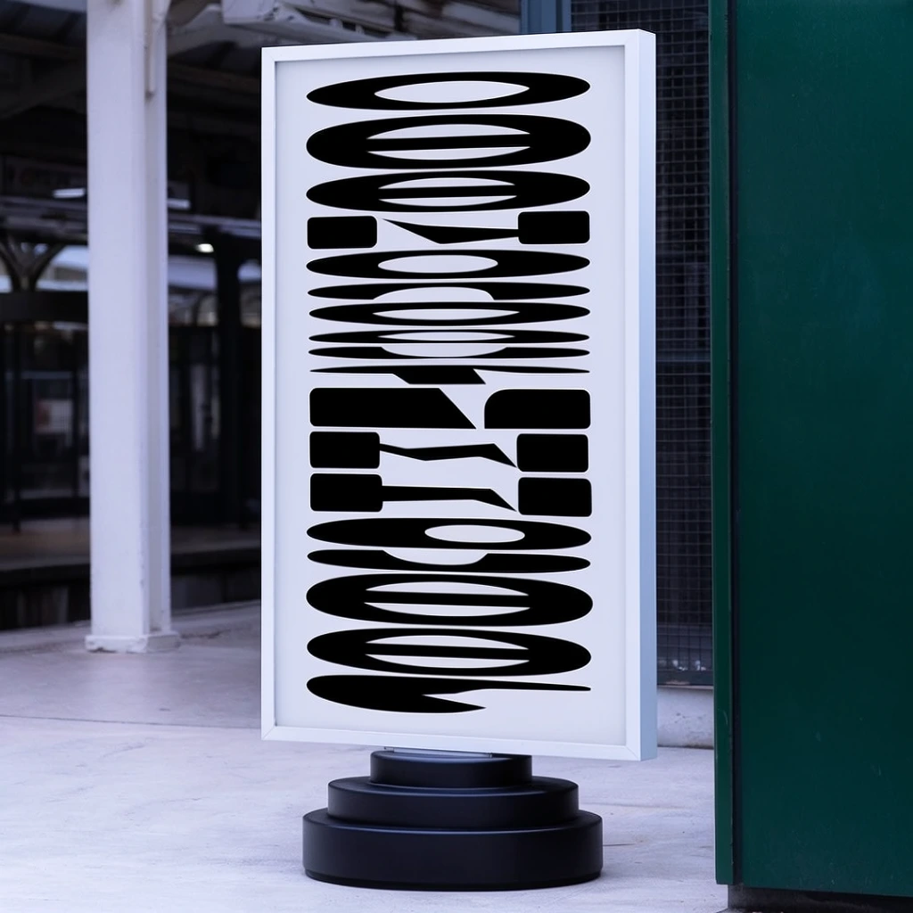 A freestanding, large vertical rectangular display board encased in a minimalist white frame stands prominently on a light concrete floor. The board is anchored by a heavy, tiered matte black circular base. The display's stark white surface is filled with dense, oversized, high-contrast black graphic shapes stacked vertically in a strict, rhythmic alignment. Adjacent to the right edge of the frame is a solid, dark green vertical structural panel. The background reveals a softly blurred urban transit or industrial environment, featuring a thick white architectural pillar, shadowed interior ceilings, and dark metal grating, all illuminated by cool, diffused daylight that highlights the shallow depth of field.