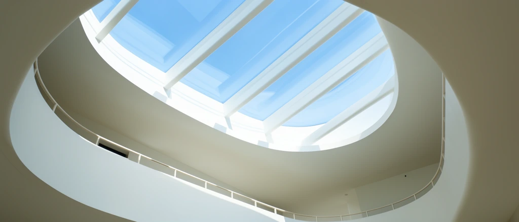 Low-angle view of an oval skylight framing a bright blue sky, smooth white curved interior walls, minimalist atrium, linear white structural beams, curved mezzanine balcony