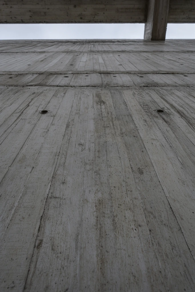 An immense, textured concrete wall dominates this extreme low-angle architectural photograph. The brutalist surface features pronounced vertical formwork striations, horizontal structural seams, and scattered circular tie-rod holes. Subtle dark streaks and weathering mottle the industrial grey expanse. At the uppermost edge, a narrow, horizontal slit of pale overcast sky is tightly framed by an overhanging concrete beam. A perpendicular structural column anchors the top right corner. The minimalist, abstract composition relies on stark geometric lines, raw materiality, and flat, diffused natural lighting to emphasize the imposing scale and weight of the structure.