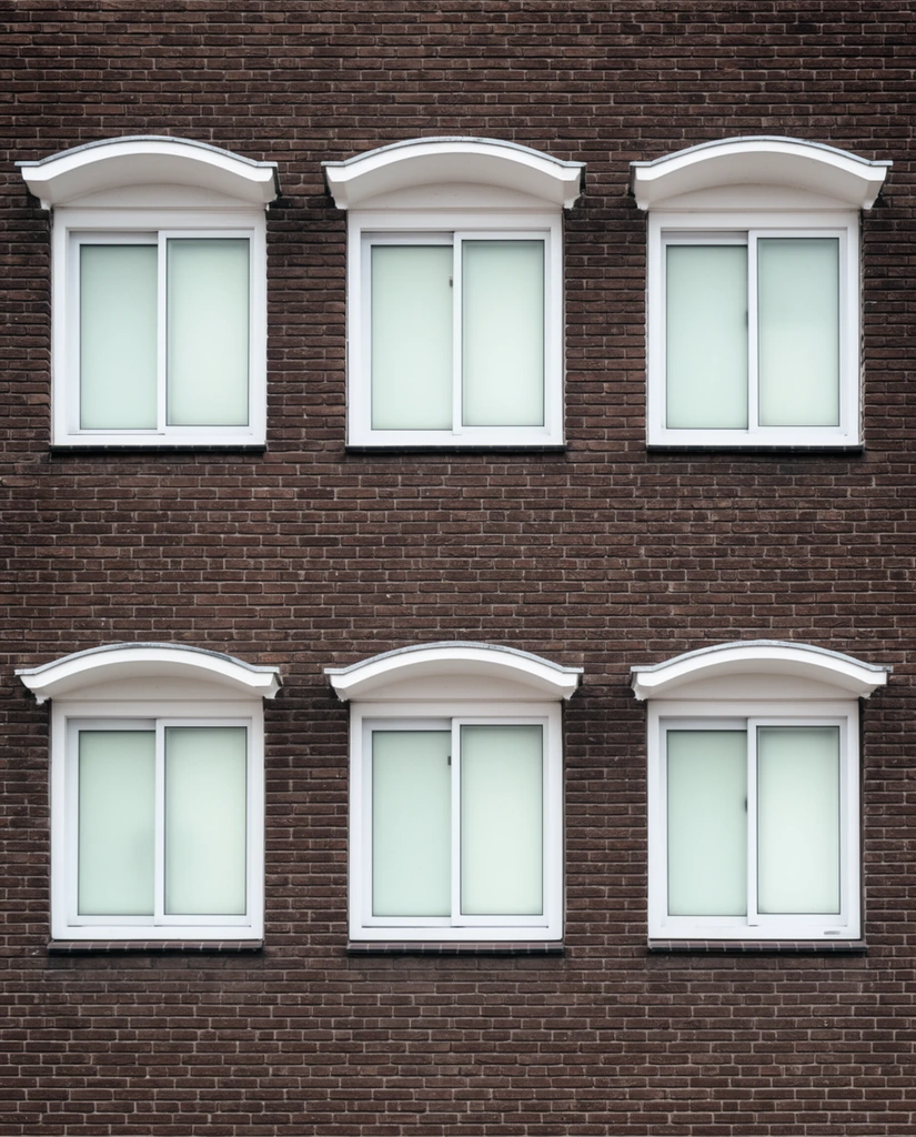 A symmetrical dark brown brick building facade features a grid of six identical windows. Arranged in two rows of three, the sliding windows have stark white frames and frosted, pale greenish-white glass panes. Distinctive white architectural hoods with shallow arches cap each window, contrasting sharply against the textured reddish-brown masonry. Captured straight-on with a flat orthographic perspective under diffused daylight, highlighting the repetitive geometric pattern and minimalist architectural design.