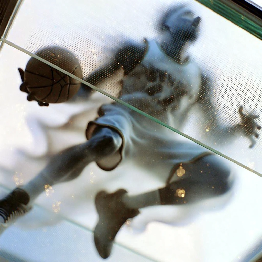 low angle perspective from directly beneath a textured frosted glass floor, dynamic silhouette of a professional basketball player suspended mid-air holding a basketball, ethereal trailing flames with a subtle glow, fine pointillist grid pattern on the translucent glass, high-key overexposed white background, soft golden bokeh light orbs, shallow depth of field, sharpest focus on areas of contact, desaturated monochromatic palette, minimalist urban aesthetic, cinematic grain