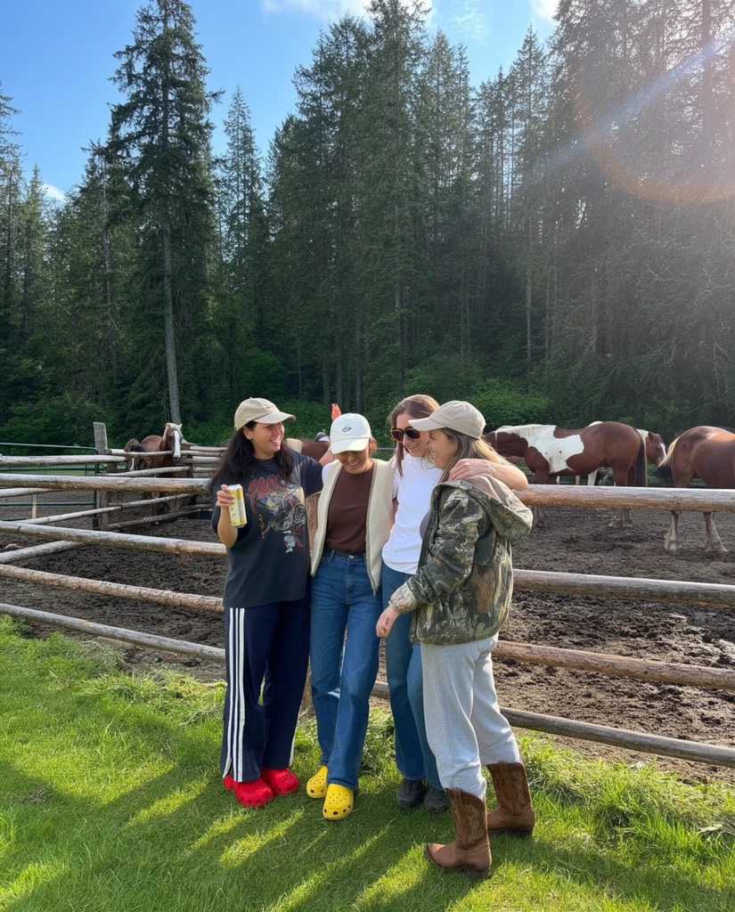 Four women stand smiling together on bright green grass beside a rustic horizontal log fence enclosing a muddy horse corral, backed by a dense forest of tall evergreen trees. The woman on the left wears a beige baseball cap, dark graphic t-shirt, dark track pants with white side stripes, and bright red clog shoes, holding a tall beverage can with one hand and slightly raising the other. Next to her, a woman in a white cap, brown t-shirt, light vest, blue jeans, and yellow clogs smiles downward. A third woman in sunglasses and a white t-shirt looks toward the center, embraced by a fourth woman wearing a beige cap, camouflage jacket, grey sweatpants, and tall brown cowboy boots. Behind the fence, several brown and white horses stand casually in the dirt. The candid, daytime snapshot features bright, natural sunlight casting long shadows, with a slight sun flare in the upper right corner against a blue sky.