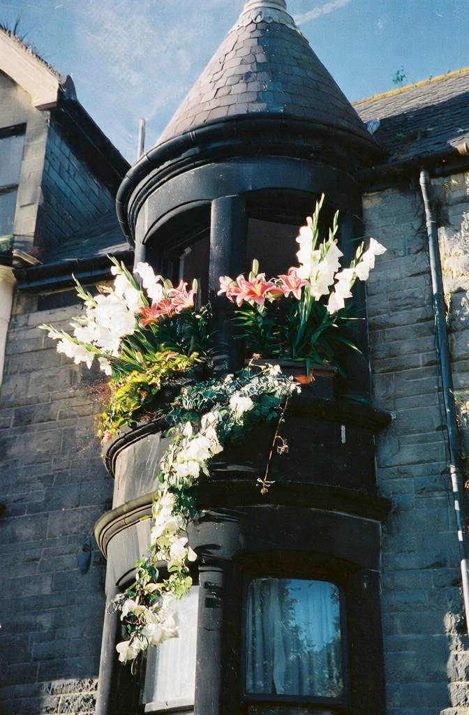 a grainy film photograph of a stone victorian house featuring a turret, pink lilies and white gladiolus overflowing from the turret windows and cascading down the facade, 35mm film aesthetic, high contrast with deep shadows, soft natural golden hour light, urban street setting, nostalgic and poetic atmosphere, vibrant yet muted color palette, snapshot style composition, rich textures, fine film grain, gritty but beautiful street photography vibe, cinematic quality.