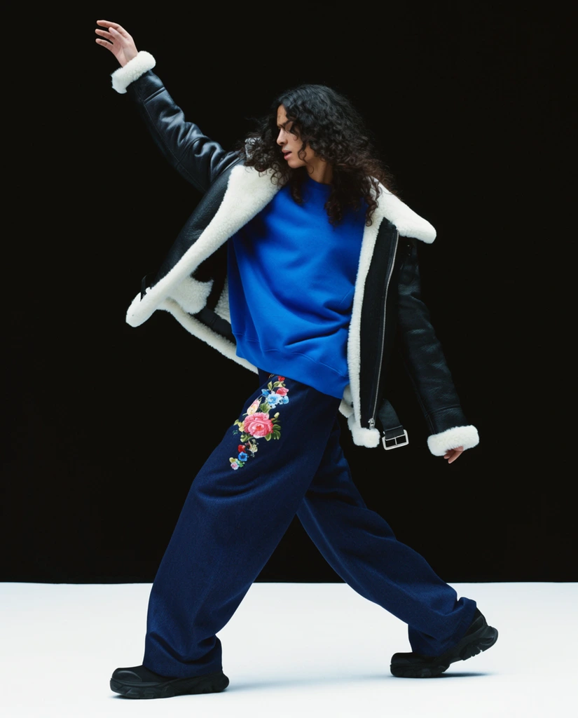 A person with long dark curly hair strikes a dynamic mid-stride pose, extending one arm high overhead. They wear a black leather aviator jacket with thick white shearling trim layered over an oversized, vibrant blue sweatshirt. Baggy dark blue trousers featuring a colorful floral motif on the left thigh drape over chunky black shoes. The minimalist environment features a stark black background abruptly meeting a bright white floor. Crisp, high-contrast studio lighting highlights the textured fabrics and fluid movement.