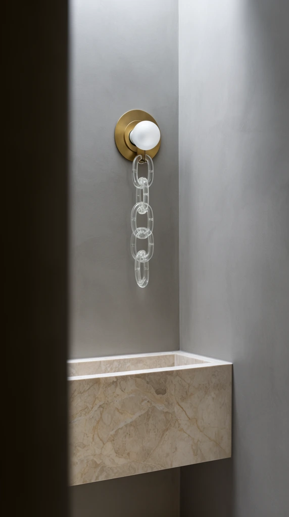 A minimalist interior alcove heavily framed by a dark, blurred vertical foreground element. Inside the recess, a floating rectangular basin of veined beige marble sits beneath a contemporary wall sconce. The fixture features a circular brass backplate, a spherical frosted glass bulb, and three oversized clear glass chain links hanging vertically. Soft, diffused overhead light washes down smooth matte grey plaster walls, highlighting crisp geometric shapes and refined material textures.