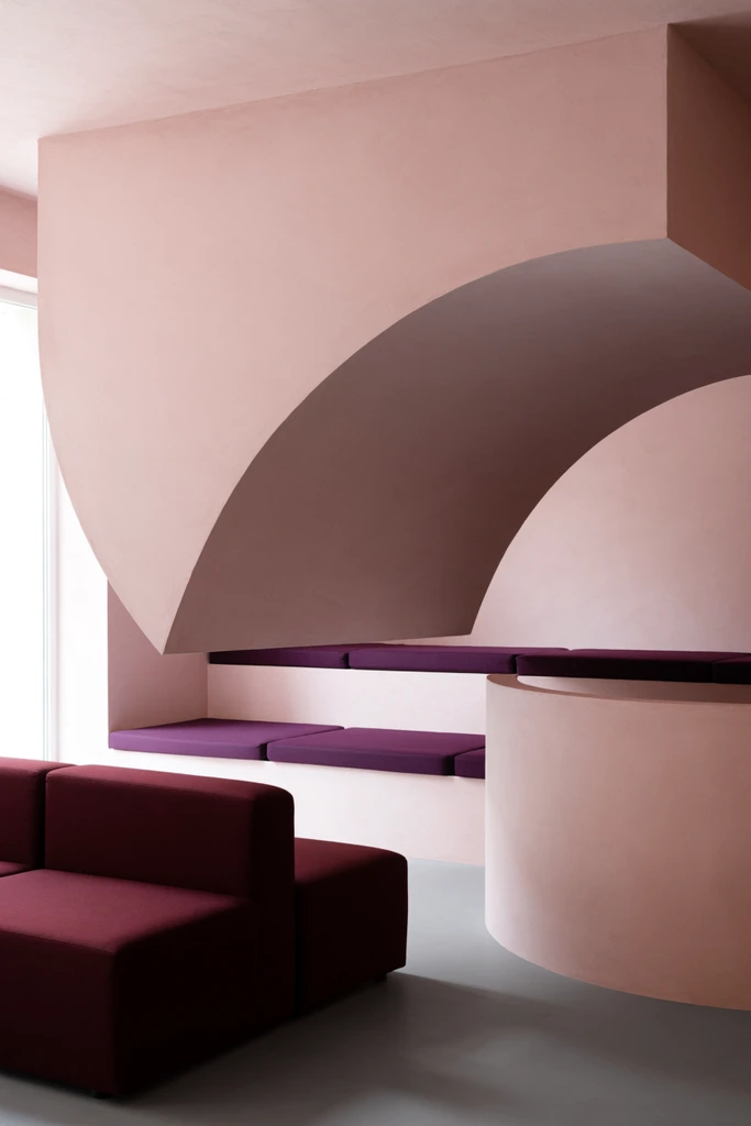 A minimalist interior dominated by a suspended dusty pink semi-circular partition. This large geometric form hangs over a built-in bench with deep plum cushions. A dark burgundy modular sofa sits in the foreground beside a curved pink half-wall on a grey floor. Soft, diffused daylight from the left highlights the smooth matte plaster finishes and reveals layered seating tiers behind the main arch. The architectural photography captures a contemporary aesthetic defined by bold overlapping geometric shapes and striking color blocking between pastel blush and saturated plum.