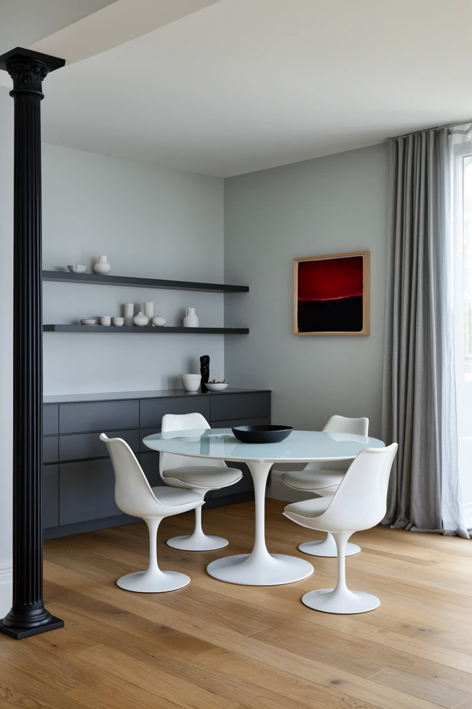 minimalist dining room, round white glass-top pedestal table, four white tulip chairs, dark grey base cabinets, floating shelves with white ceramics, pale grey walls, black fluted column, window with grey drapes, oak floors, framed abstract crimson and black painting, soft natural daylight, interior photography