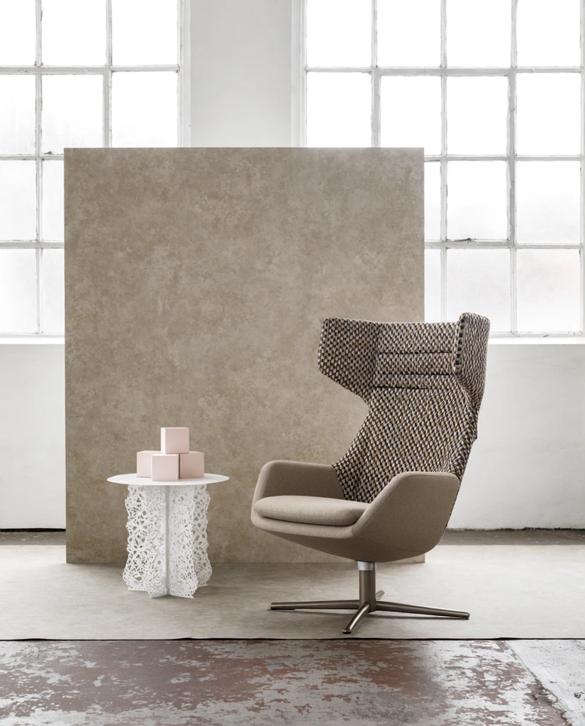 modern armchair with high woven patterned backrest and beige seat, metal swivel base, white lace-like side table holding pale pink geometric blocks, mottled beige textured backdrop panel, industrial loft environment with large white grid windows, distressed concrete floor, soft diffused daylight, minimalist interior photography, muted earthy tones