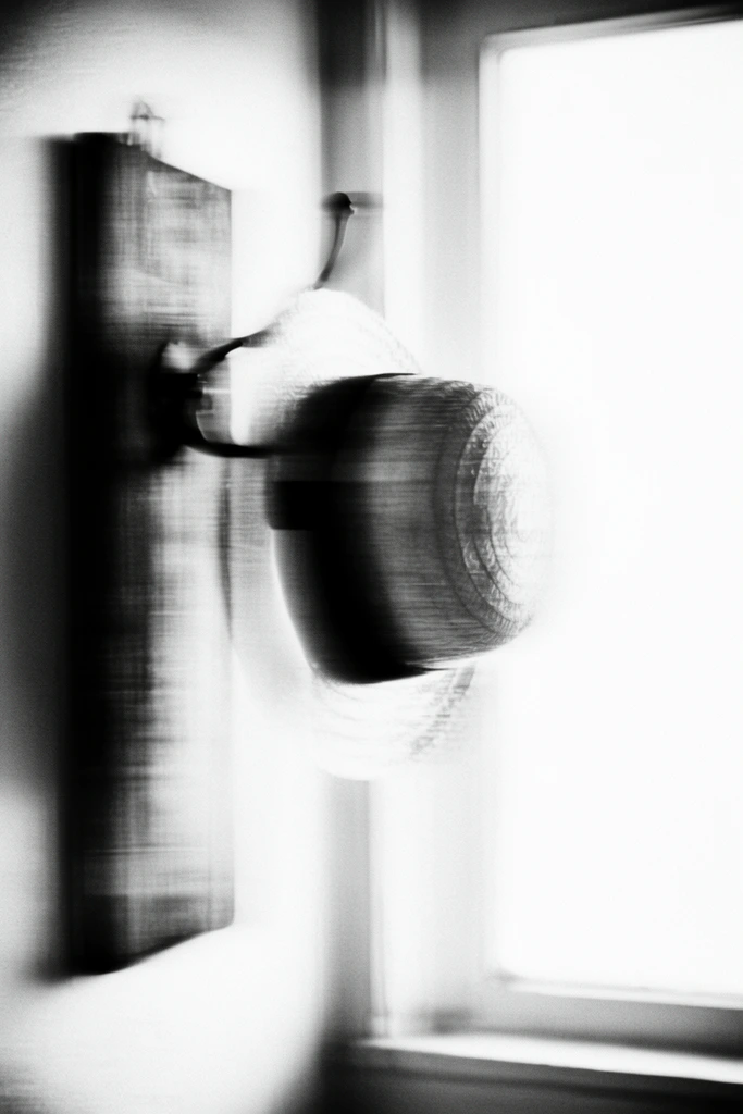 A heavily blurred, high-contrast black-and-white composition of a straw hat with a band hanging on a hook attached to a vertical wooden board on a wall beside a window. The hat's circular form is severely distorted, its edges melting into smudged, indistinct transitions. A dark band on the hat creates a thick, smeared horizontal streak across the crown. The hook and the vertical wooden board manifest as dense, shadowy vertical blocks, their textures dominated by intense horizontal motion blur. Adjacent to the board, the window forms a massive, overexposed expanse of stark white, casting intense, diffused light that washes out a large portion of the frame. The entire scene is rendered in a monochromatic palette featuring crushed blacks and blown-out whites, mimicking the effect of abstract, slow-shutter photography or an image captured through heavily frosted glass. Coarse film grain, stark chiaroscuro, and sweeping horizontal smearing transform the physical objects into soft, continuous visual gradients.