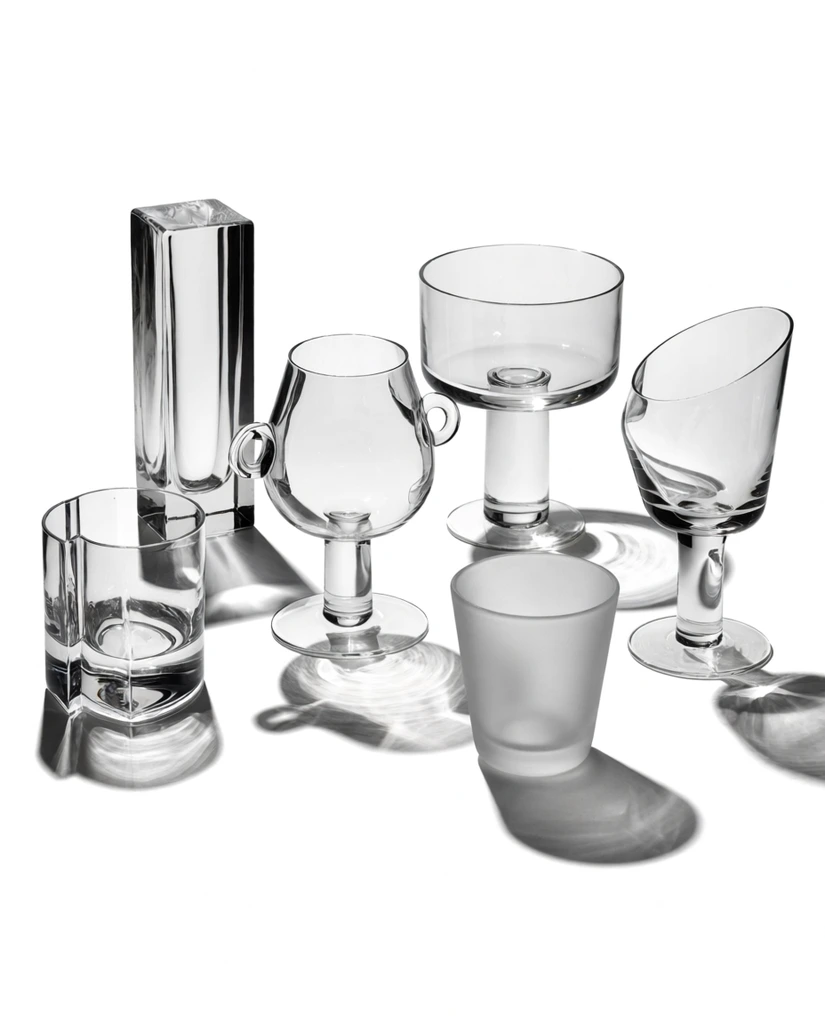 six distinct clear glass vessels on a pure white surface, tall rectangular prism, folded cylindrical tumbler, bulbous stemmed glass with side loops, wide pedestal bowl, frosted cup, asymmetrical stemmed glass, monochrome still life photography, strong directional studio lighting, brilliant optical refractions, sharp shadows, complex geometric caustics