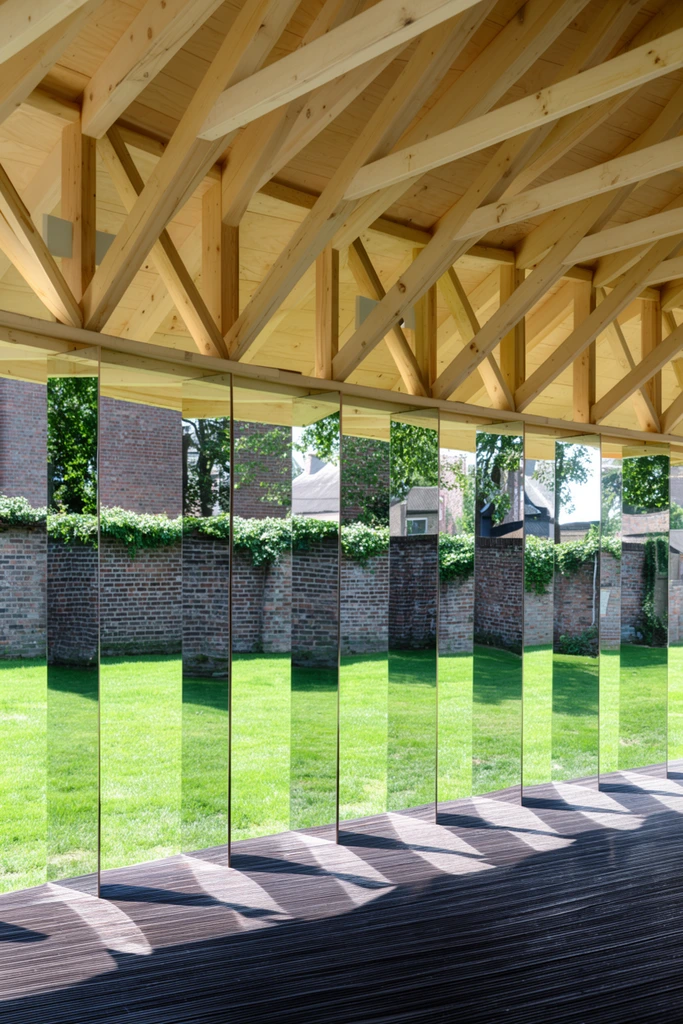 modern open-air pavilion, exposed light wood roof trusses, vertical mirrored panels reflecting a green lawn and brick wall, dark slatted wood floor, bright sunlight, architectural photography