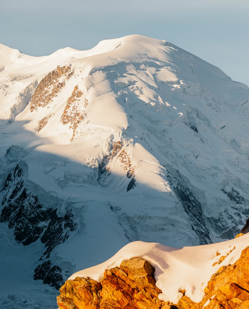 A massive snow-covered mountain peak dominates the frame under a pale blue sky. The upper face is bathed in crisp sunlight, highlighting intricate textures of glacial ice, deep crevasses, and sharp ridges. A dramatic diagonal shadow cuts across the left side, exposing dark, jagged rock walls beneath the snow. In the immediate foreground, a rugged outcrop of orange-brown stone capped with smooth snow catches intense golden-hour light, creating a stark, warm contrast against the immense, cool-toned alpine face. The telephoto composition emphasizes the sheer scale and verticality of the rugged landscape.