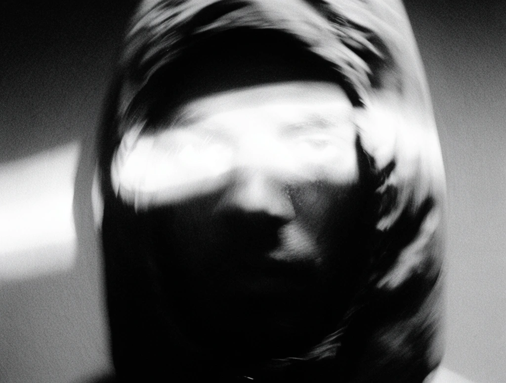 A close-up portrait of a face enclosed in a hood with a horizontal light beam obscuring the eyes. The heavy fabric of the hood and the lower facial contours merge into deep, crushed black shadows, forming a dense, graphic silhouette. A harsh, overexposed streak of pure white light cuts across the upper face, flooding the eye line with intensely blooming highlights. The image is executed in extreme high-contrast black and white, characterized by severe motion blur that smears the forms into dynamic, sweeping trails. Coarse film grain, heavy optical distortion, and a swirling soft focus saturate the frame, replicating a raw, lo-fi analog aesthetic. The stark collision of blinding, halating whites and absolute blacks creates a gritty, textured atmosphere dominated entirely by stark extremes of luminance.