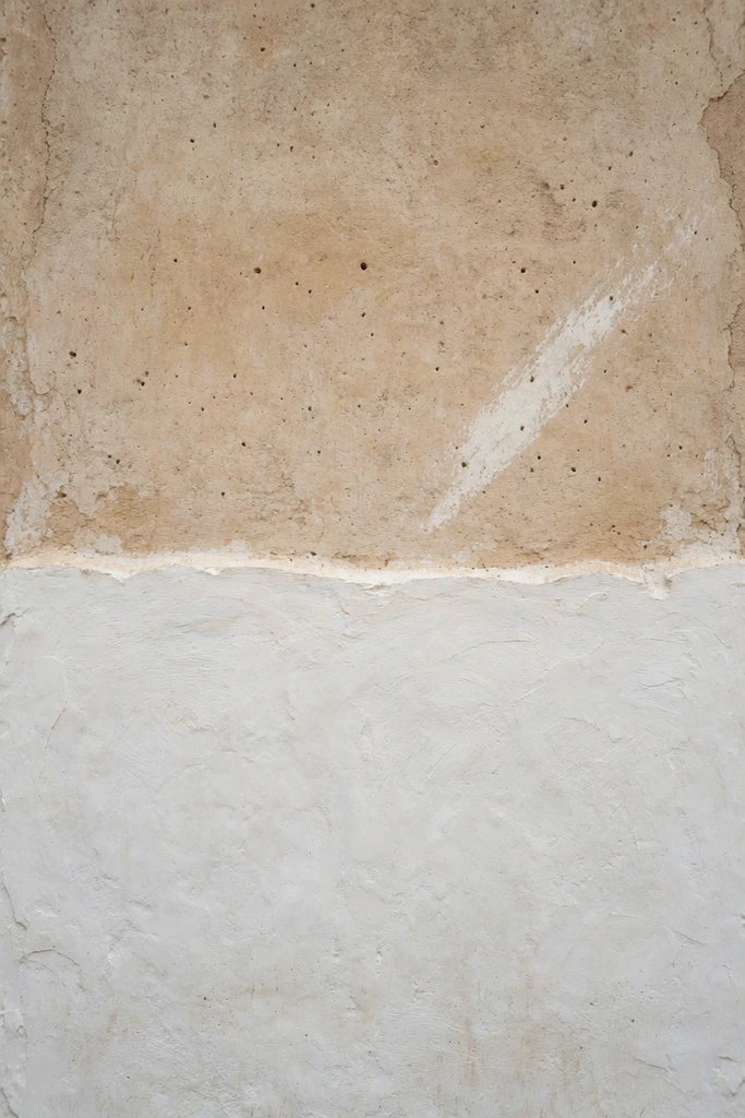 A minimalist, two-toned textured wall divided horizontally by an uneven border. The upper section consists of aged, warm tan plaster with small pockmarks and a distinct diagonal white smear. The lower half features a thick, roughly applied layer of bright white plaster. Soft, flat lighting emphasizes the raw, tactile architectural materials and contrasting wabi-sabi aesthetic.