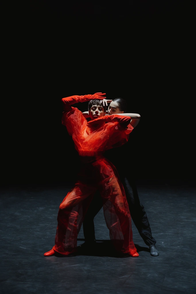 Two dancers perform in a pitch-black void. The foreground figure wears a voluminous sheer red outfit and gloves, posing dynamically with arms framing their face. Behind, a dancer in dark clothing places a pale hand on the first's head. High-contrast spotlighting isolates the subjects on a smooth dark floor, creating deep shadows in a cinematic, center-framed wide shot.