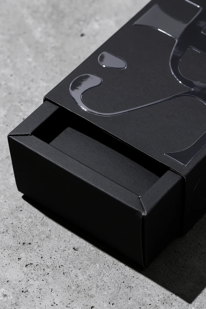 A partially open matte black rigid cardboard box rests on a textured light gray concrete surface. The packaging features a sliding inner tray made of thick dark paperboard with crisp, sharp edges. Glossy, raised clear-coated abstract motifs catch the light across the lid, contrasting with the smooth matte finish. Strong angled lighting from the top left casts deep, harsh shadows across the porous stone background. The minimalist macro product photography emphasizes the interplay of premium paper textures and industrial stone in a stark monochromatic palette.